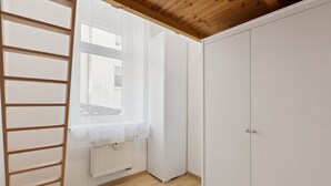 Una habitación con techo de madera, paredes blancas, un radiador, un ventanal con cortinas traslúcidas y un armario blanco.