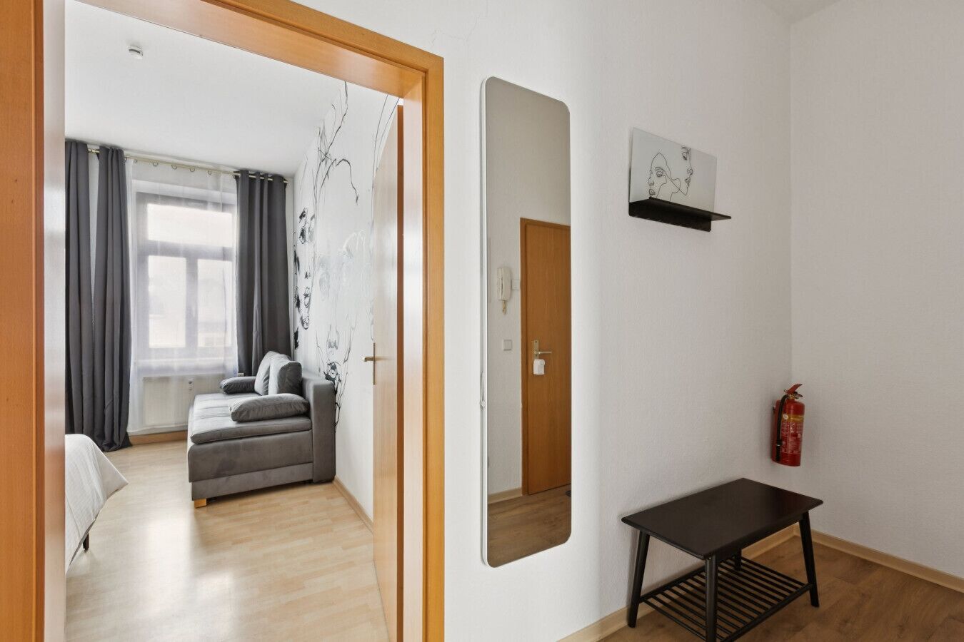 A room with a bed, a sofa, a small table, a mirror, a fire extinguisher, and a window with curtains.