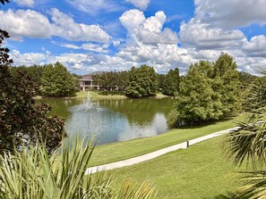 A tranquil pond with a fountain, surrounded by well-maintained grass and trees, with a building in the background. - Disney Area 3BR Condo Pickleball, Pools & Free WiFi (Davenport)