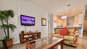 Ein Wohnzimmer mit Flachbildfernseher, einem Glastisch, einem blumigen Sofa und einem Holzkonsolentisch mit Büchern.