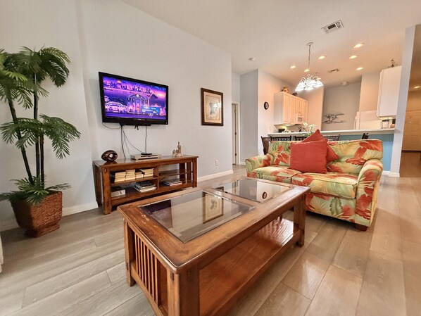 A living room with a flat-screen TV, a glass-top coffee table, a floral sofa, and a wooden console table with books. - Relax in Paradise! Spacious Bahama Bay Condo Near Disney (Davenport FL)