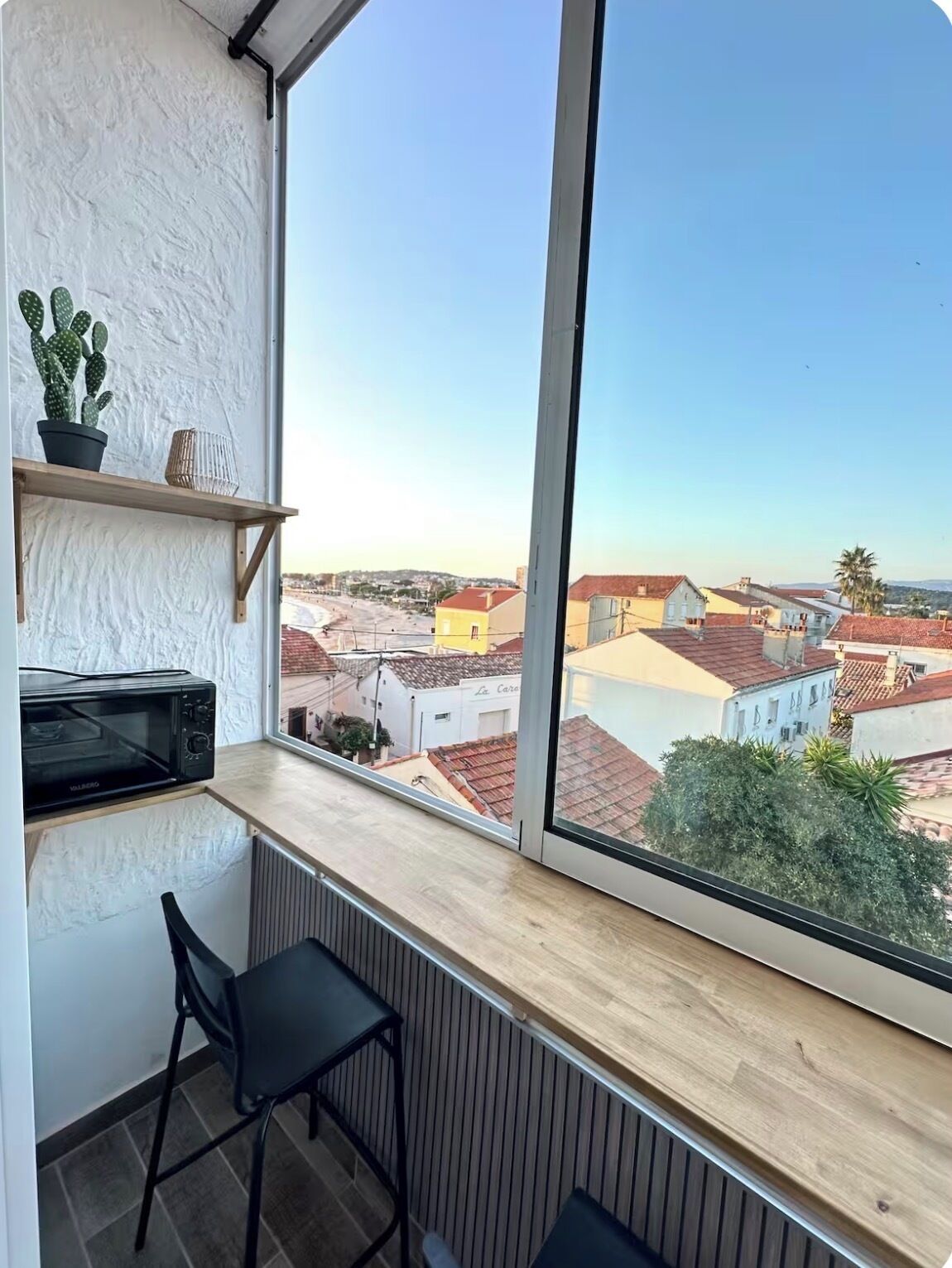 A room with a large window, a wooden shelf with a cactus and a bowl, a black chair, and a microwave.