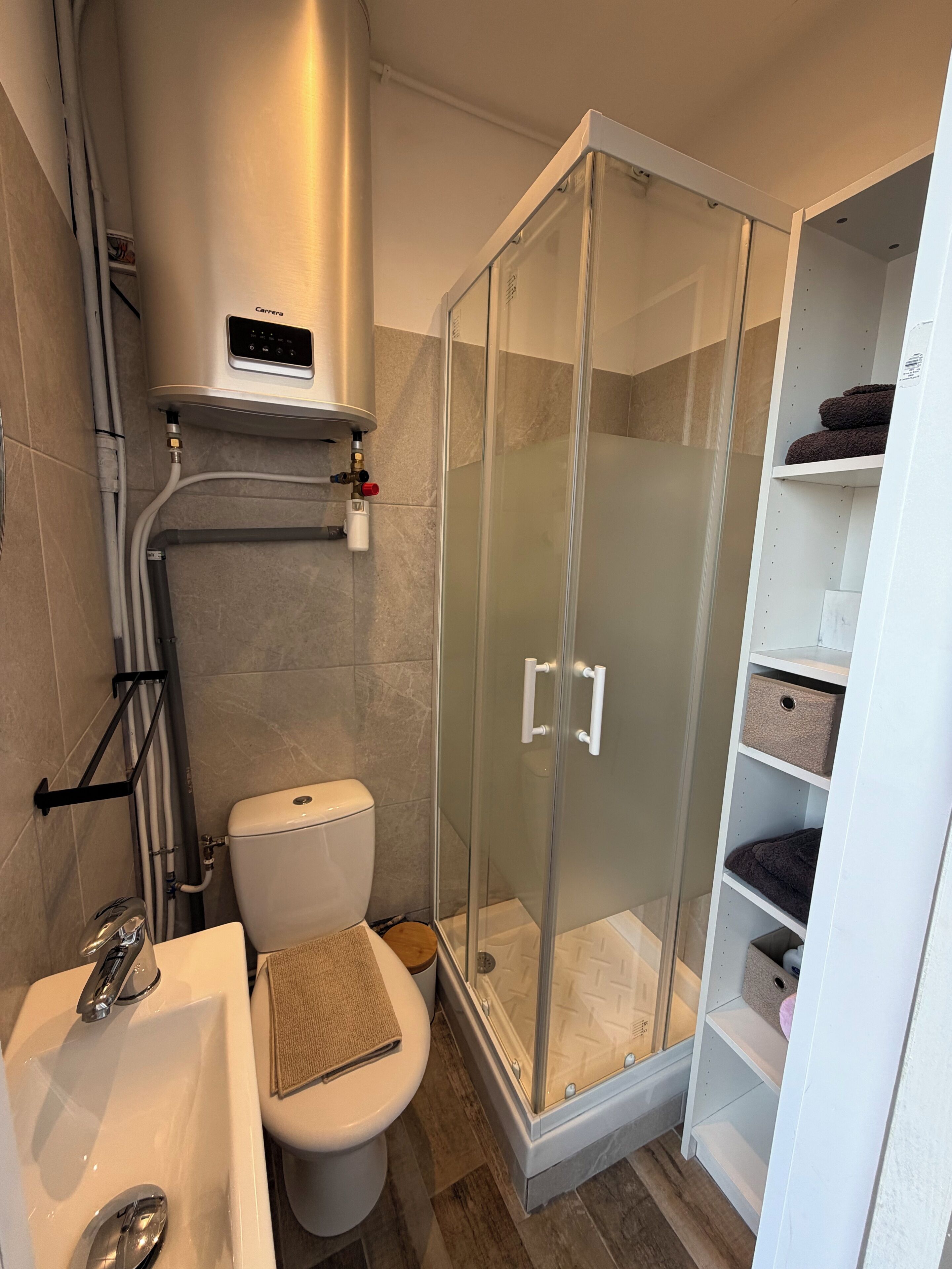 Une salle de bain comprenant des toilettes, un lavabo, un espace douche et des étagères de rangement.