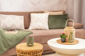 En hyggelig stue med en sofa, puder, et lille rundt bord med et stearinlys, en fletkurv og en potteplante.