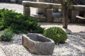 Uma bacia de pedra em um jardim com chão de cascalho, cercada por vegetação e estruturas de pedra.