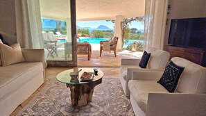 Una sala de estar con una mesa de centro de vidrio, sofás color beige y vistas a una piscina y montañas.