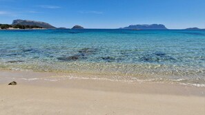 Una playa con agua cristalina, orilla arenosa y montañas a lo lejos.
