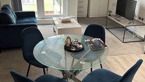 Ein modernes Wohnzimmer mit einem Glastisch, blauen Stühlen, einer Couch, einem Fernseher und einer weißen Kommode.
