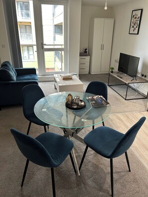 Un soggiorno moderno con un tavolo da pranzo in vetro, sedie blu, un divano, una TV e un mobiletto bianco.