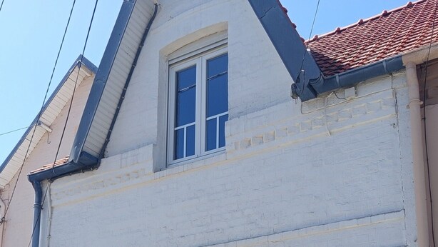 Un edificio de dos plantas con fachada blanca y contraventanas azules, que presenta un pequeño ventanal con cortina de encaje.