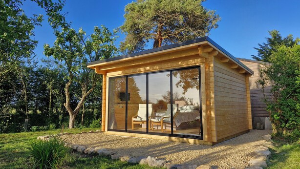 Una cabaña de madera con grandes puertas de cristal, un patio con sillas y un camino de grava.
