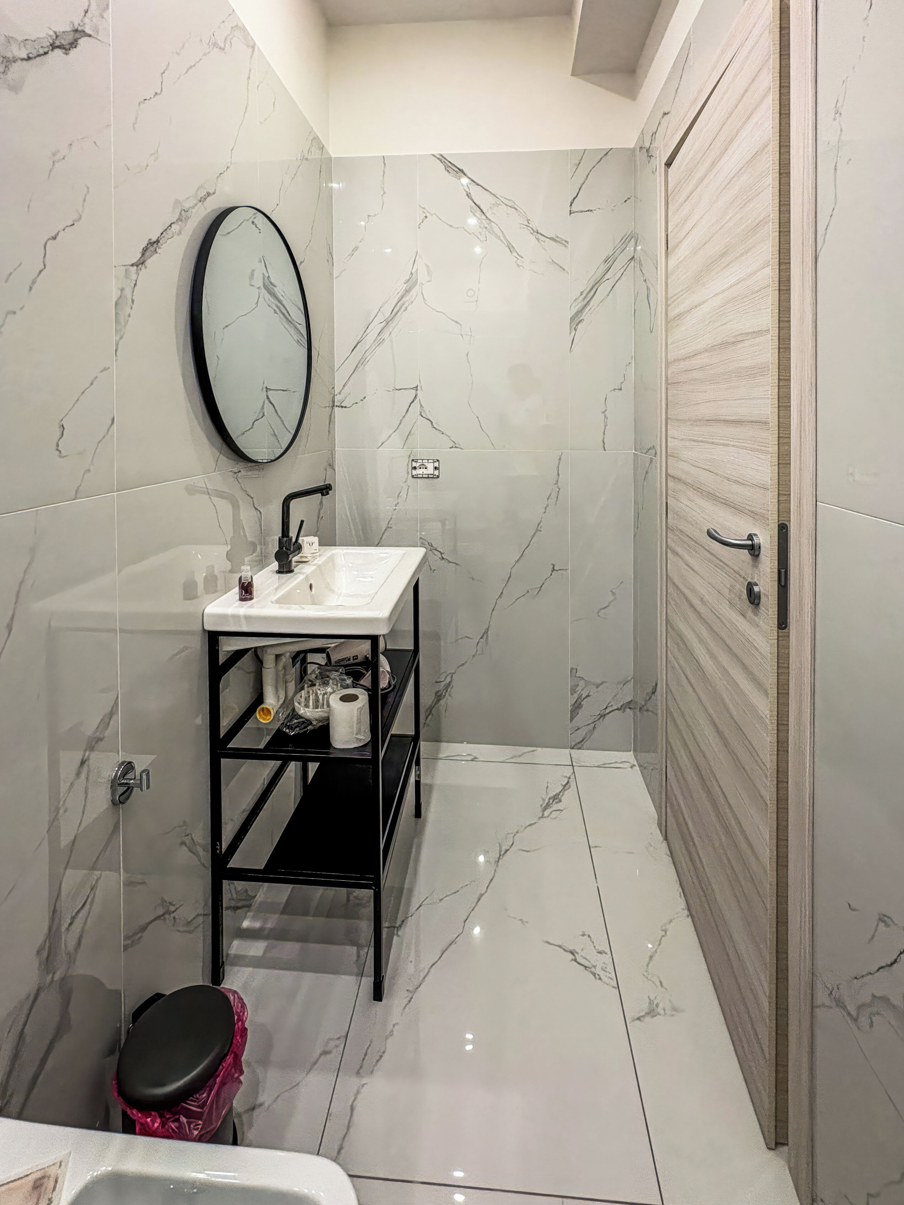 A bathroom with a white sink, mirror, and black shelf; a toilet with a pink trash bin; and a closed door.