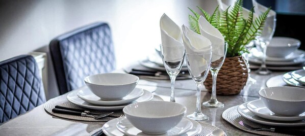 Una mesa de comedor dispuesta con cuencos, platos y cubiertos blancos, un vaso de agua y una planta de helecho.