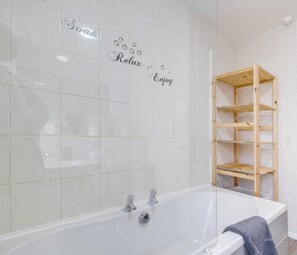 Un baño con una bañera blanca, una estantería de madera y azulejos decorativos que dicen 'Soak', 'Relax' y 'Enjoy'.