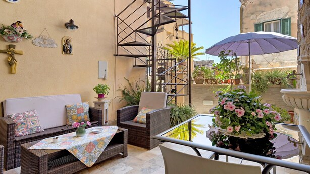 Ein Balkon mit Korbmöbeln, einer Metall-Spiralkonstruktion als Treppe, Topfpflanzen und einem violetten Sonnenschirm.