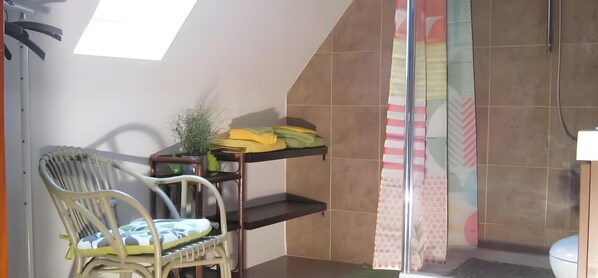 Une salle de bain avec une douche, des toilettes, une chaise et une Ă©tagĂšre oĂč sont pliĂ©es des serviettes.