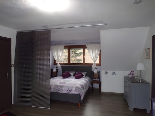 A bedroom with a bed, a nightstand, a dresser, a window with curtains, and a door.