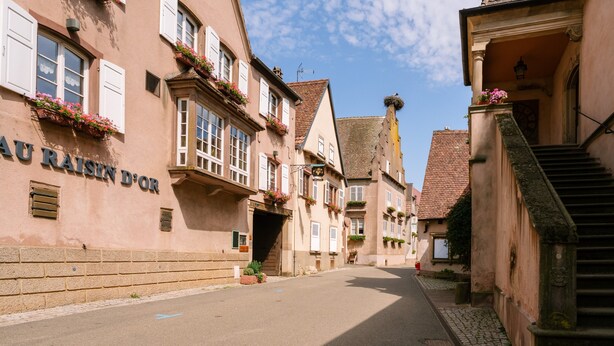 Eine enge Straße gesäumt von traditionellen europäischen Gebäuden, bewölkter Himmel, und ein Schild mit der Aufschrift 'AU RAISIN D'OR'.