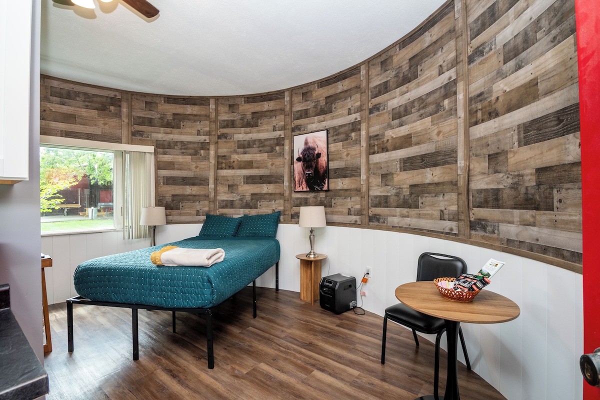 A room with a curved wooden wall, a bed with teal bedding, a small table with a basket, and a lamp.