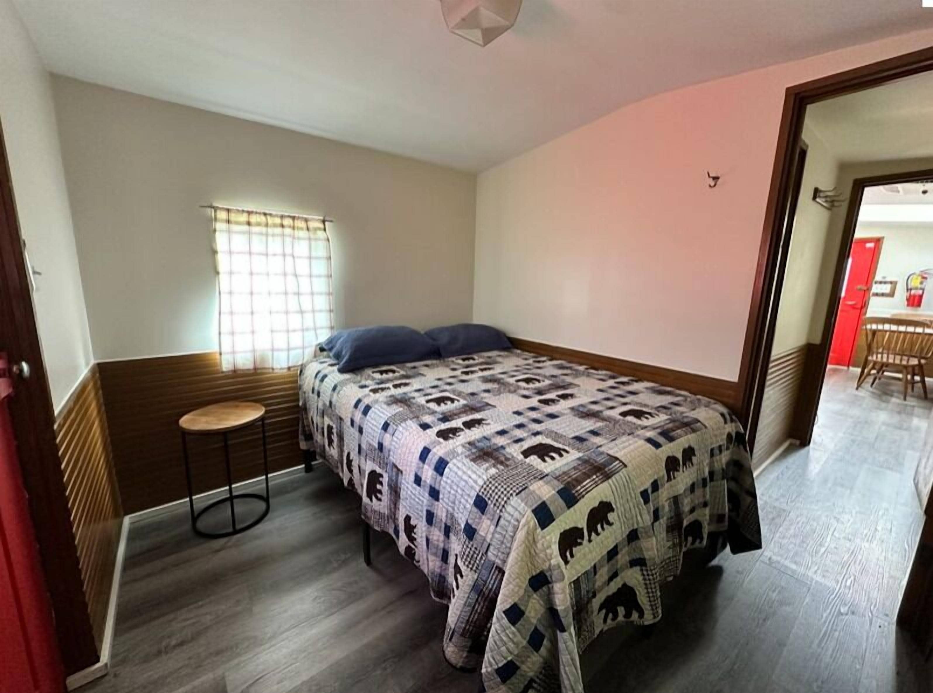A bedroom with a bed, a small stool, a window with curtains, and a door leading to another room.