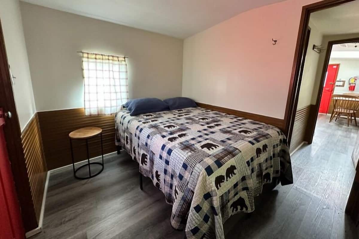 Un dormitorio con cama, un taburete pequeño, una ventana con cortinas y una puerta que da a otra habitación.
