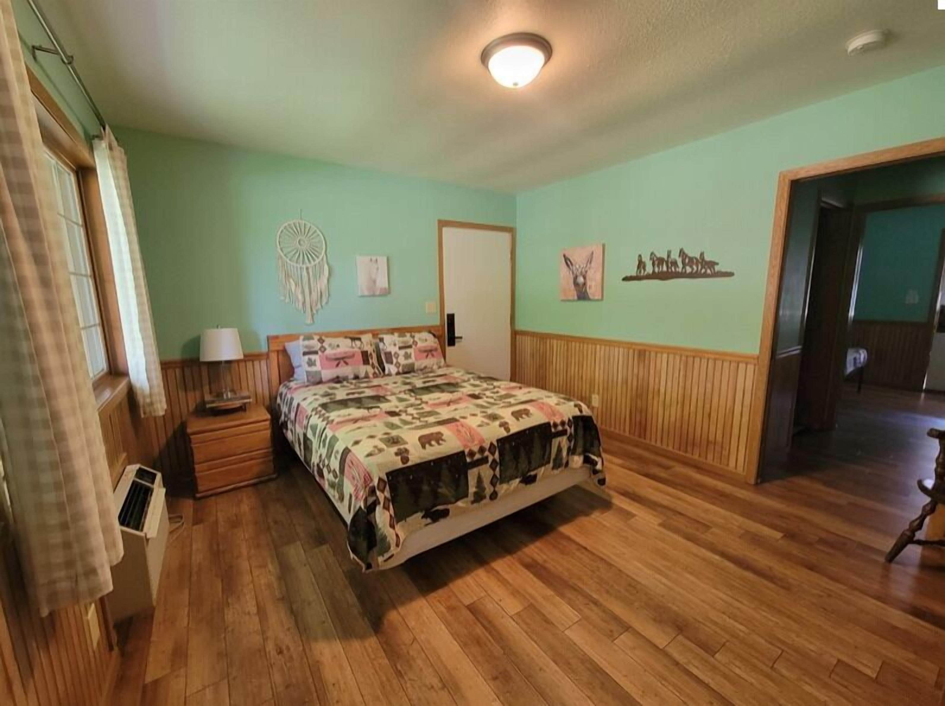 A bedroom with a wooden floor, a bed with a patterned bedspread, a bedside table with a lamp, a wall decoration, and a door leading to another room.