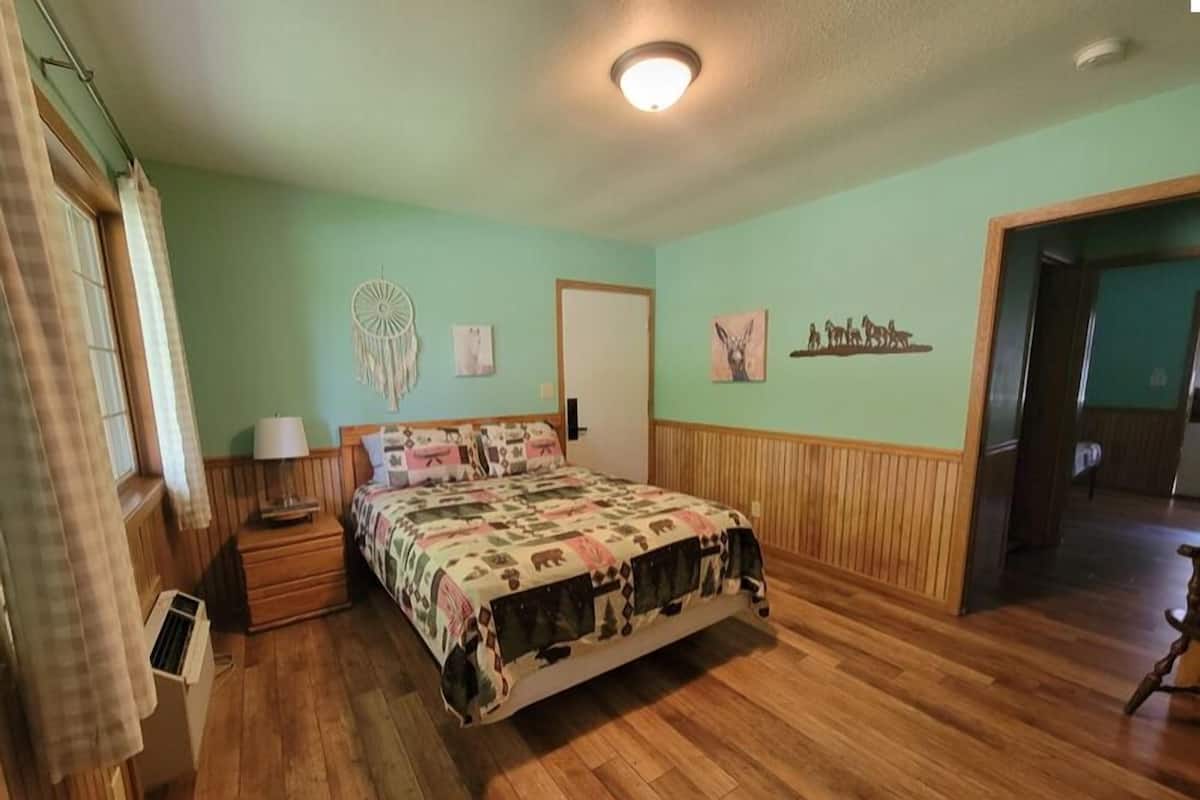 Un dormitorio con piso de madera, una cama con cubrecama estampado, una mesita de noche con lámpara, una decoración en la pared y una puerta que da a otra habitación.