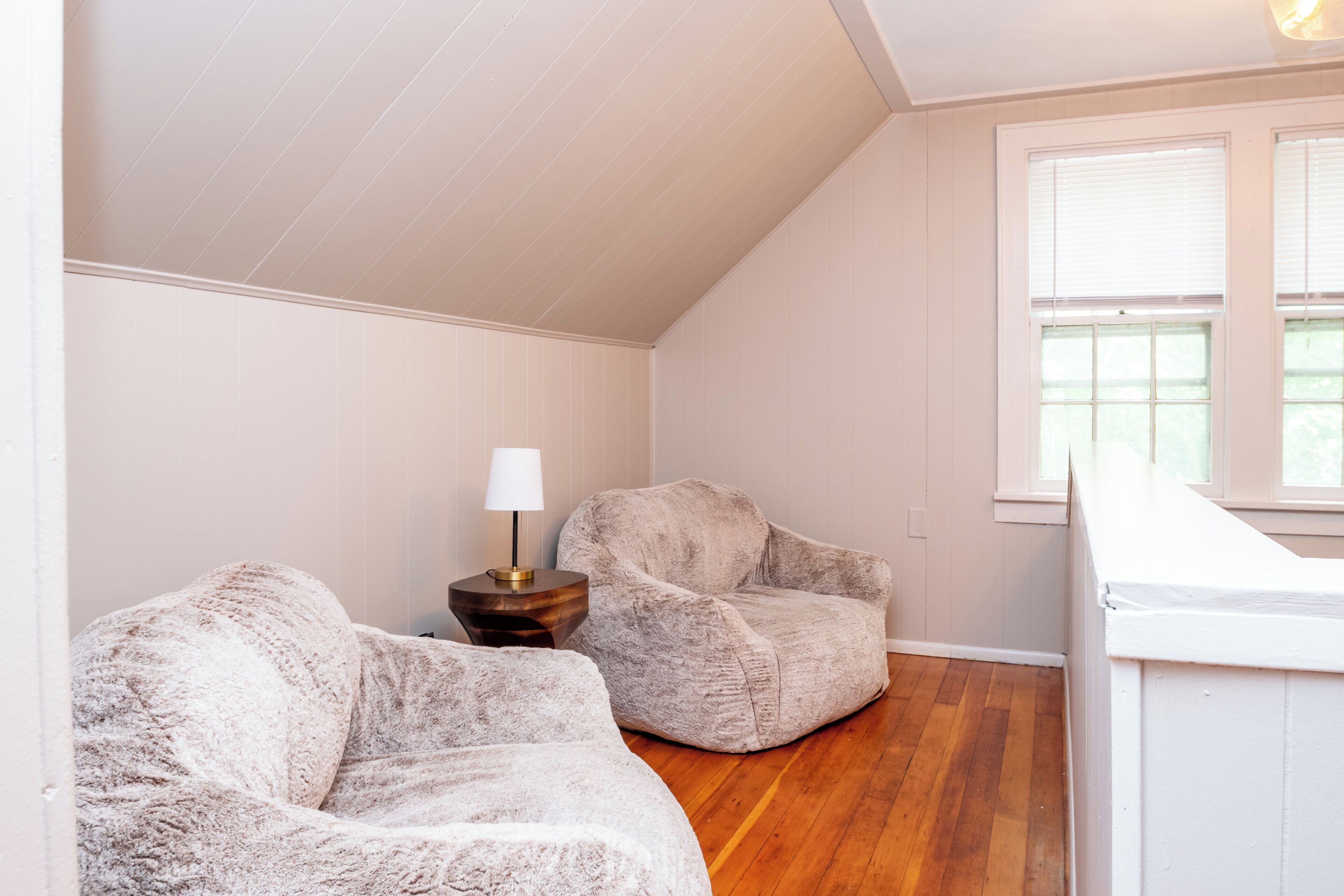 Una habitación con techo inclinado, dos sillones mullidos, una pequeña mesita de madera con una lámpara y una ventana con persianas.