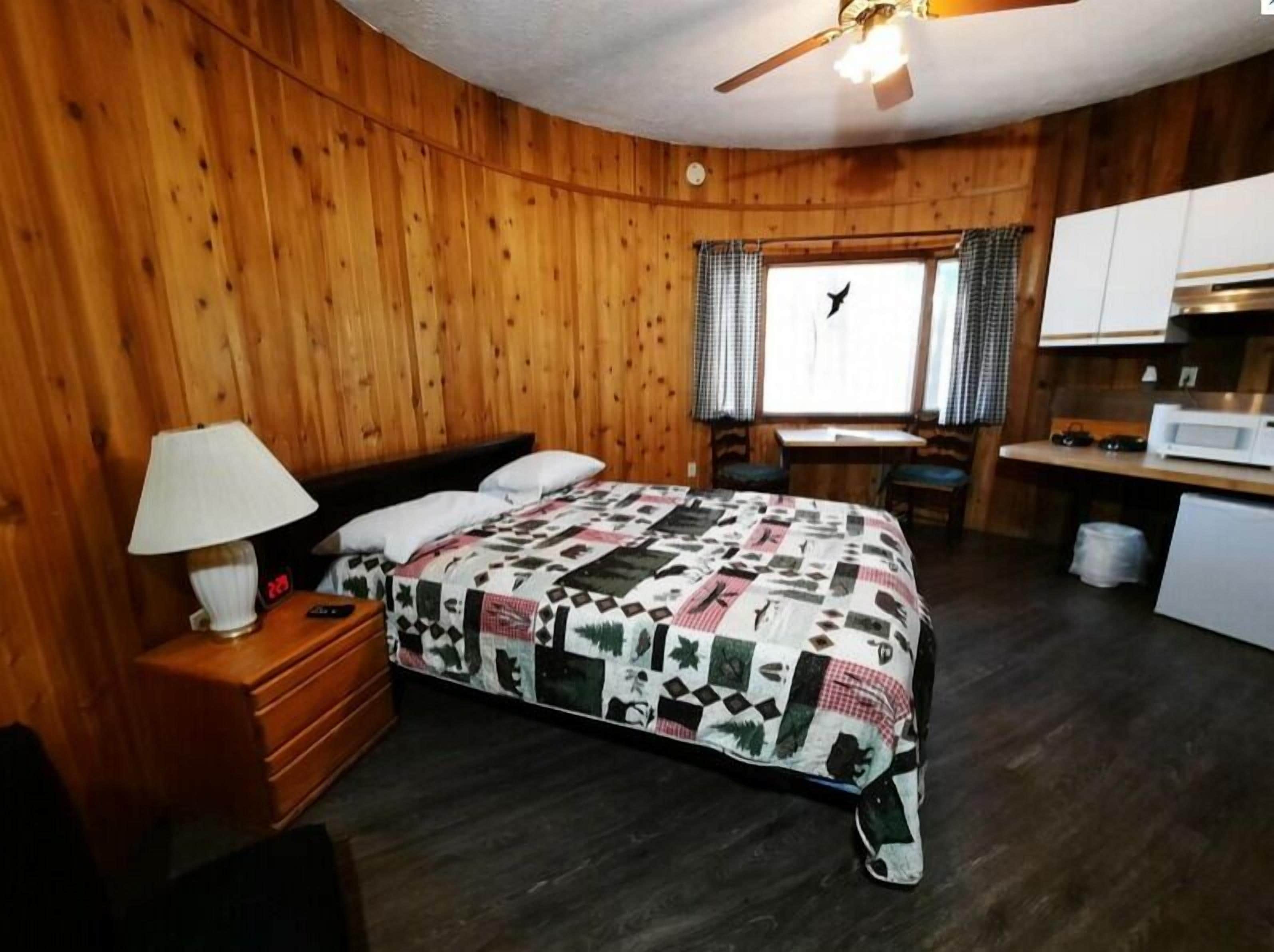 A wooden-paneled room with a bed, a desk, a chair, a lamp, and a window with curtains.