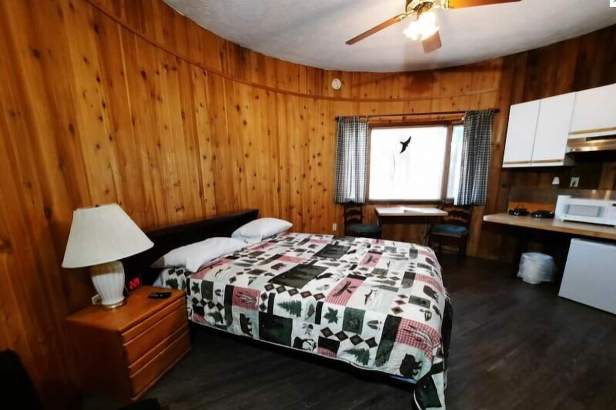 Una habitación con paneles de madera, una cama, un escritorio, una silla, una lámpara y una ventana con cortinas.