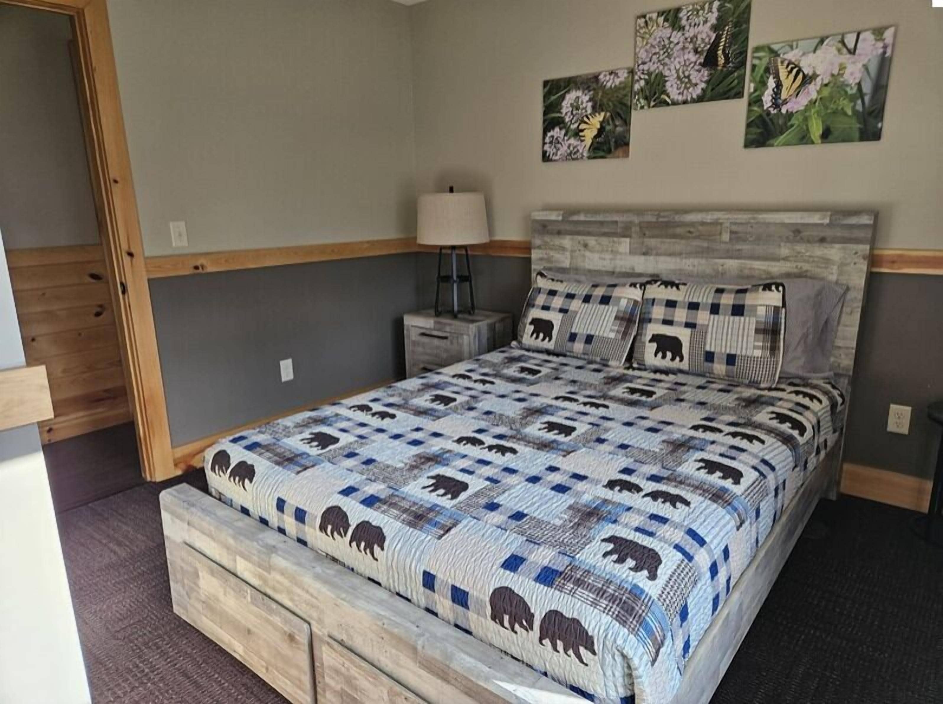 A bedroom with a wooden bed frame, a quilt with animal patterns, a nightstand, a lamp, and wall art.