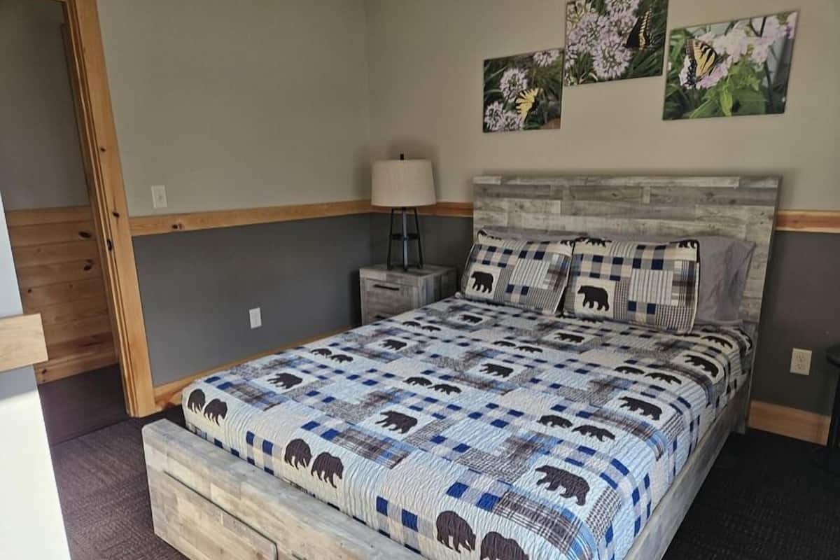 Un dormitorio con estructura de cama de madera, colcha con estampados de animales, mesita de noche, lámpara y decoración en la pared.