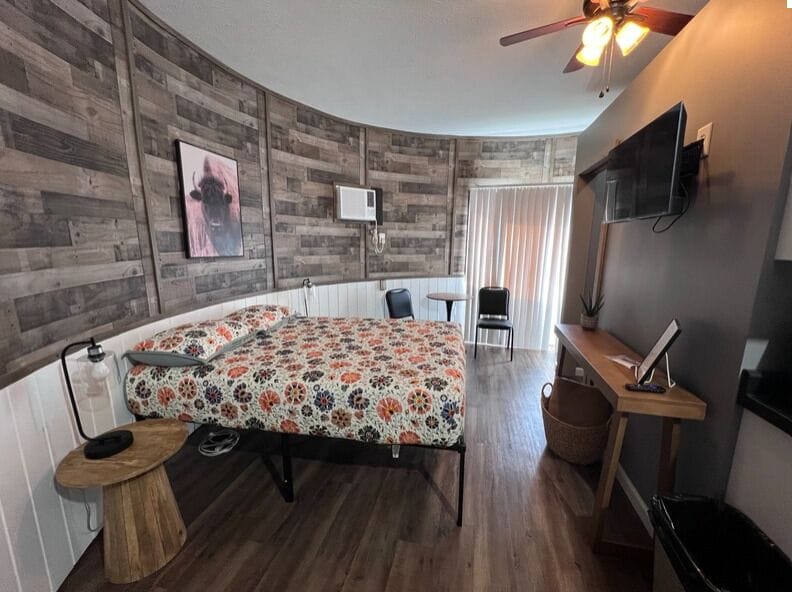 A bedroom with a curved wooden wall, a bed with a floral bedspread, a desk with a chair, a television, and a ceiling fan.