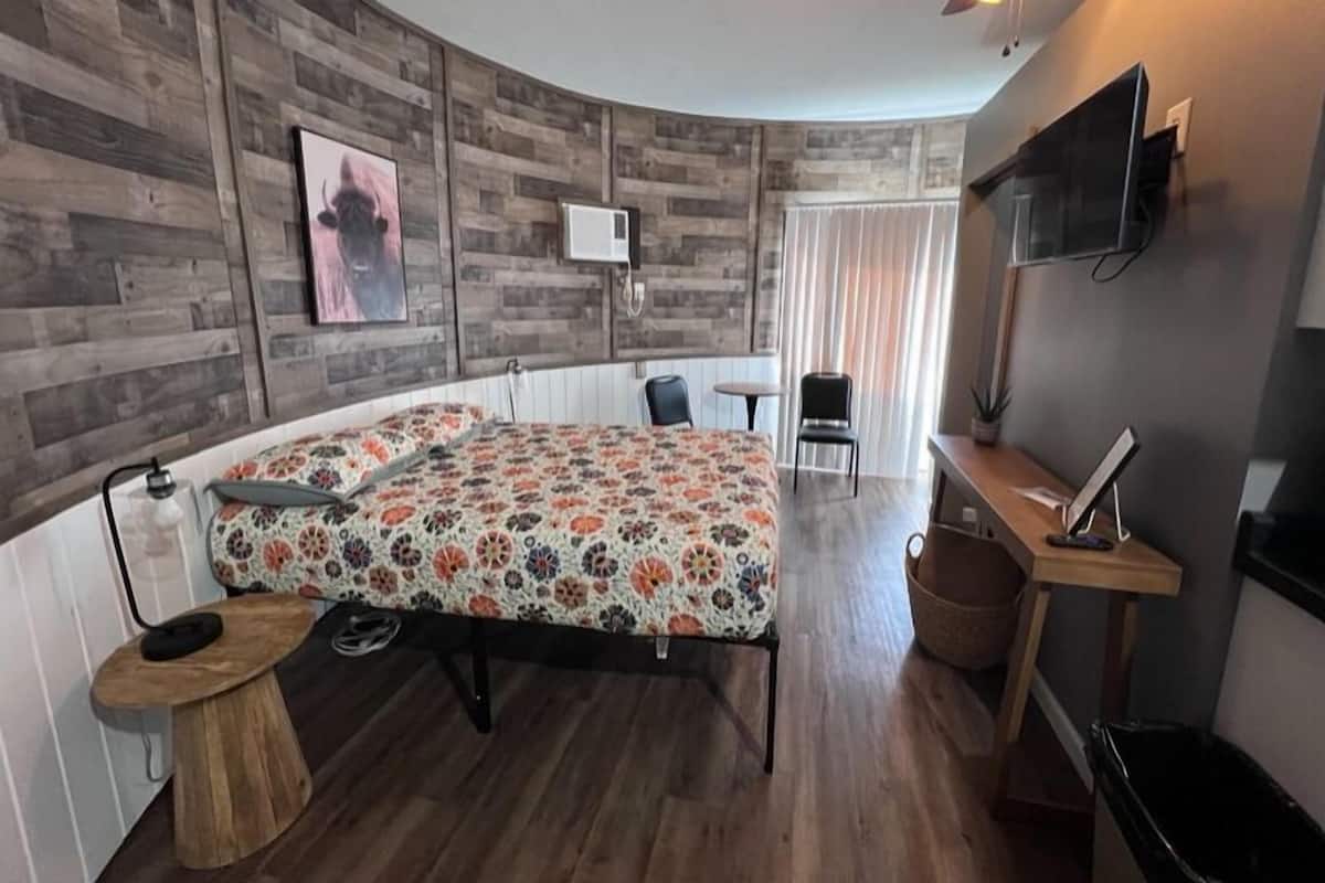 Un dormitorio con una pared de madera curva, una cama con colcha estampada, un escritorio con silla, un televisor y un ventilador de techo.