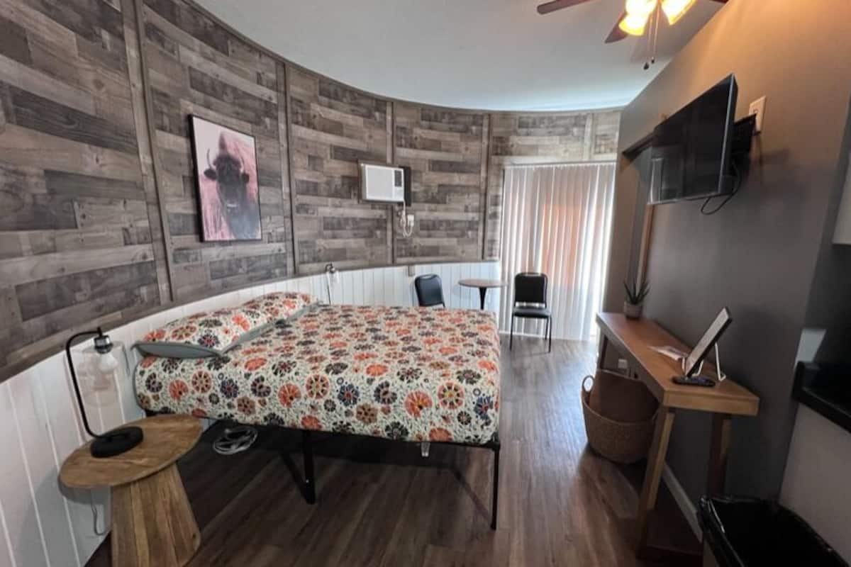 Un dormitorio con una pared de madera curva, una cama con colcha estampada, un escritorio con silla, un televisor y un ventilador de techo.