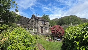 A stone house with a white fence, surrounded by lush greenery and a mountain backdrop. - Over Brow Cottage (Coniston)