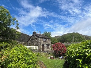 Ein aus Stein gebautes Haus mit weißem Zaun, umgeben von üppiger Vegetation und einem Berg im Hintergrund.