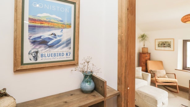 A framed poster of a speedboat on Coniston Water, a wooden bench, a vase with dried plants, a white sofa, and a rocking chair in a living room. - Over Brow Cottage (Coniston)