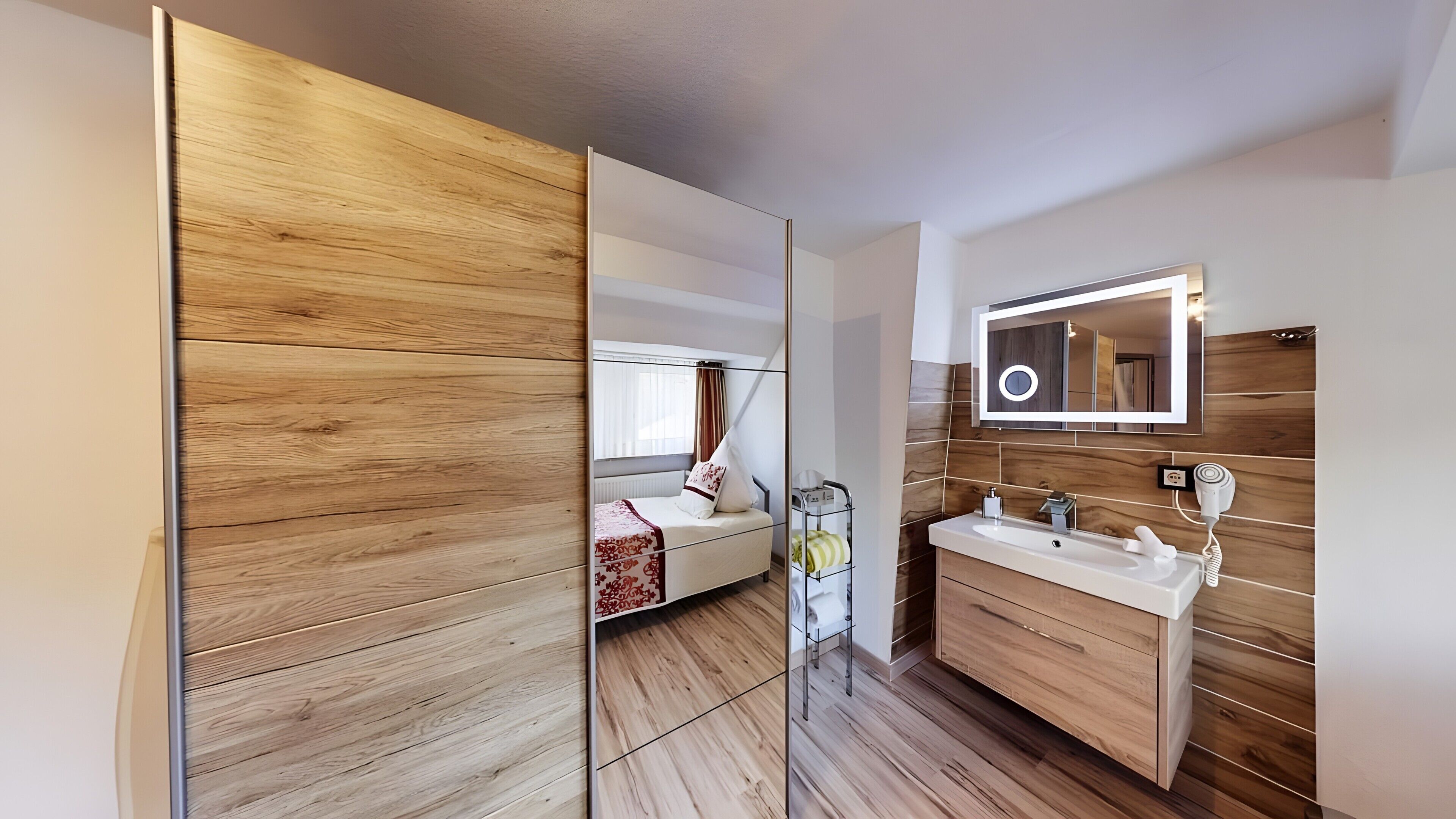 A modern bathroom with a wooden vanity, a mirror with lights, and a bed in the background.