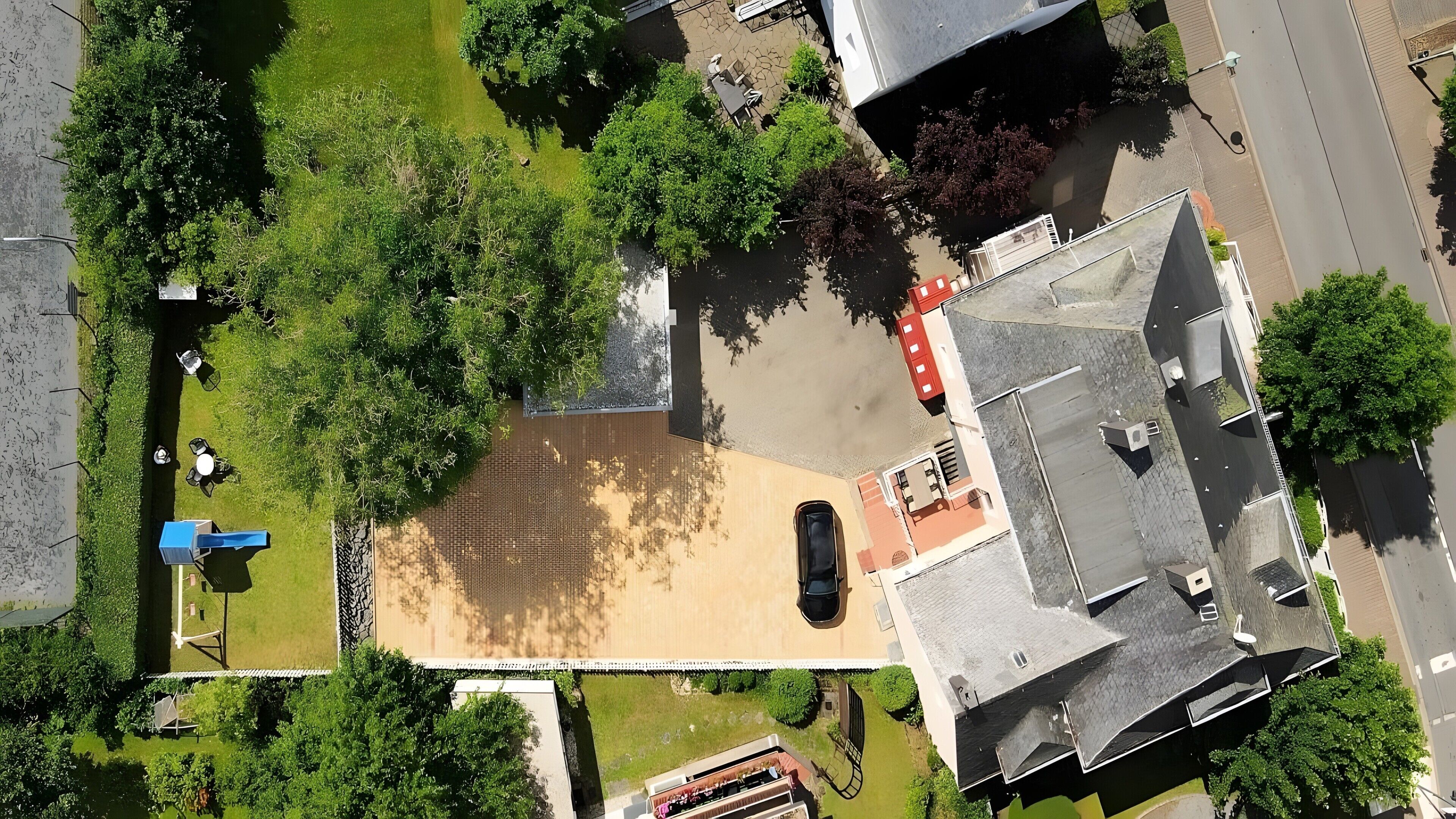 An aerial view of a property with a paved courtyard, surrounding greenery, and a building with a sloped roof.