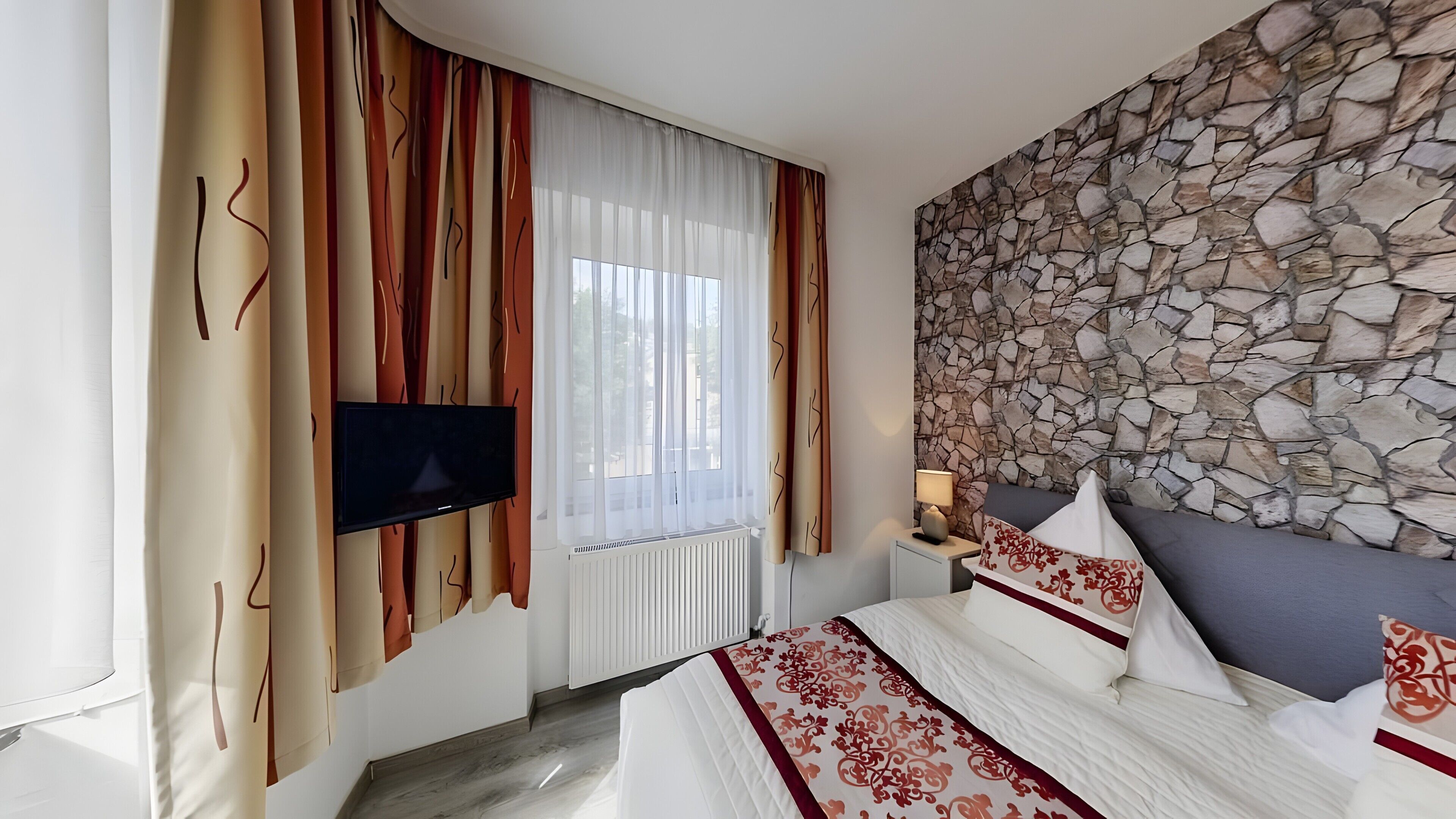 Hotelkamer met een stenen muur, een bed met beddengoed met een patroon, een flatscreen televisie en een raam met dunne gordijnen.