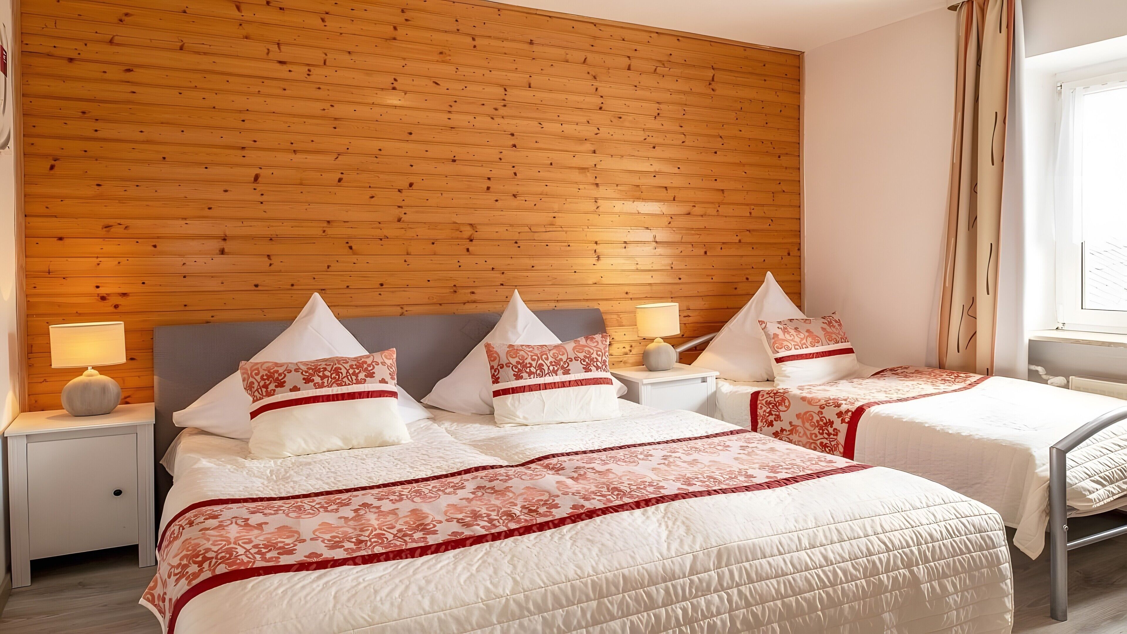A hotel room with a wooden panel wall, two beds with matching headboards, bedside tables with lamps, and a window with curtains.