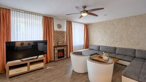 A modern living room with a flat-screen TV, a stone fireplace, a curved sofa, and a ceiling fan. - City Holiday Home Daun in Daun – for 14 Guests (210 m²) (Daun)
