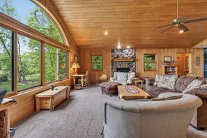 Interior acogedor de una cabaña con chimenea, muebles de madera y ventanas amplias que ofrecen vistas al exterior.