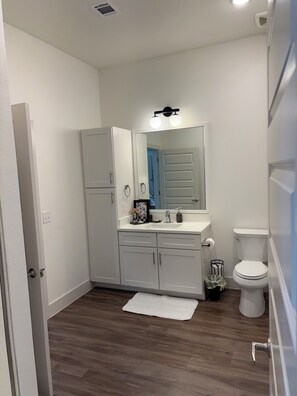 Un bagno con lavandino bianco, specchio e WC, pavimento in legno e tappeto bianco.