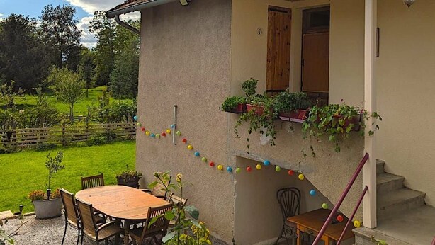 Una casa con tetto in tegole, un portico coperto, un'area pranzo con tavolo e sedie, e una scala che conduce all'ingresso.