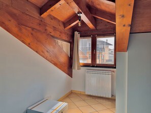 Ein Zimmer mit schrägverdeckter Decke, Holzbalken, Heizkörper und einem Fenster mit Blick auf Nachbargebäude.