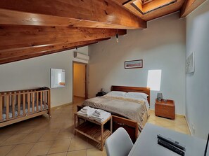 Een slaapkamer met een houten plafond, een bed, een ledikant, een bureau met stoel en een nachtkastje met lamp.