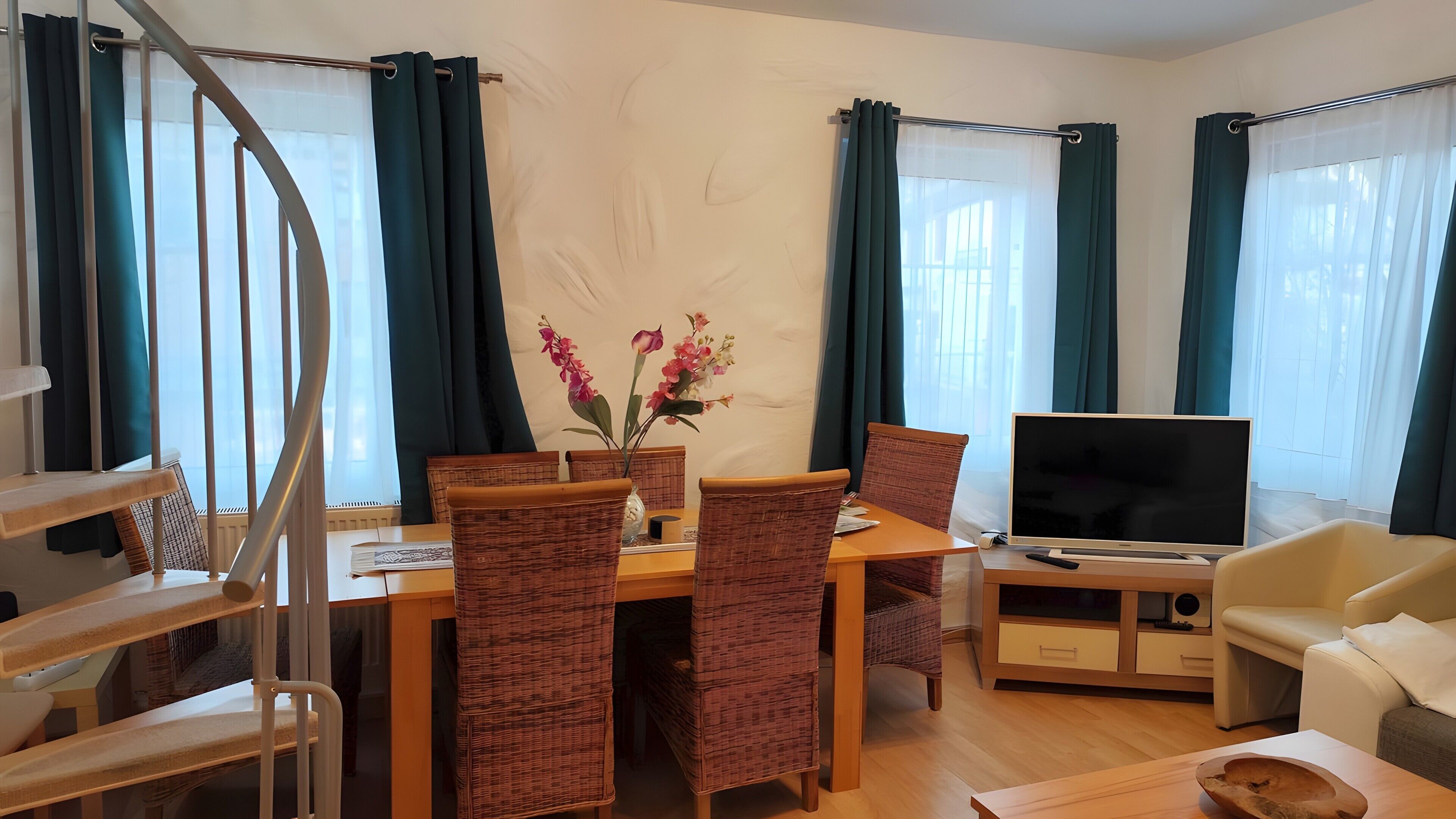 A modern living room with a dining table, wicker chairs, a flat-screen TV, a sofa, and a staircase.
