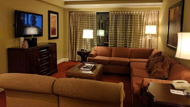 Ett hotellrum med en brun soffa, en tv, ett skrivbord med en lampa och ett fönster med gardiner.